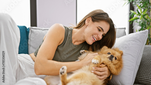 Confident young hispanic woman enjoying a fun laugh, hugging her pet dog while sitting joyfully on her home sofa with a beautiful smile.