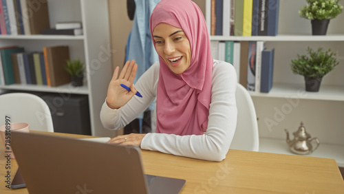 Smiling woman in hijab using laptop at home, waving during a video call in a modern interior. © Krakenimages.com