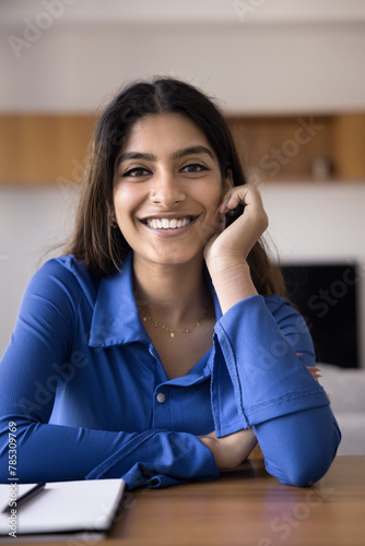 Cheerful beautiful 20s Indian girl student sitting at home workplace table, leaning chin on hand, looking at camera with toothy smile. Happy young freelancer woman vertical head shot portrait