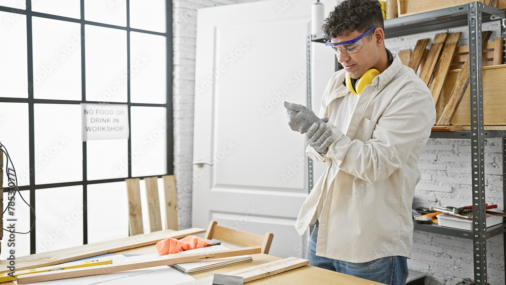 Αυτοκόλλητο A young man examines his wrist in pain in a well-lit carpentry workshop filled with wood and tools