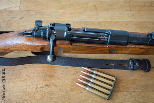 Detail of an ammunition clip with 5 30-06 caliber rifle bullets next to a Mauser-type bolt-action military rifle on a wooden table