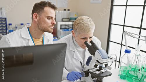 A woman and man in lab coats work together, analyzing samples with a microscope in an indoor laboratory.