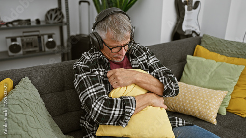 A grey-haired man listens to music with headphones, hugging a yellow pillow on a grey sofa in a cozy home interior.