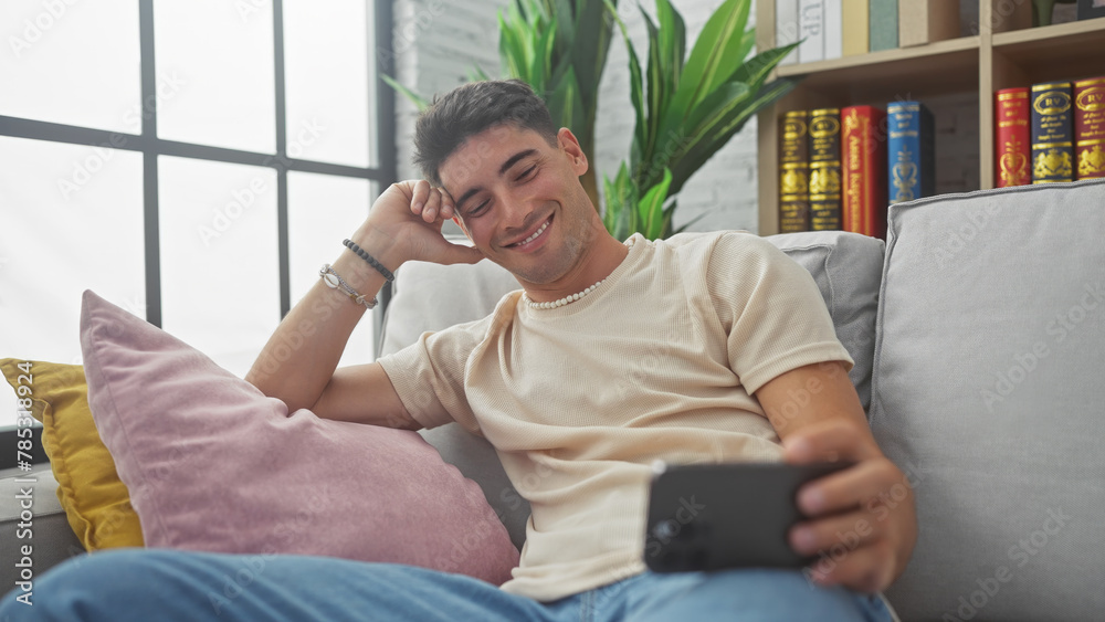 Smiling young hispanic man lounging on sofa with smartphone at home Smiling young hispanic man lounging on sofa with smartphone at home