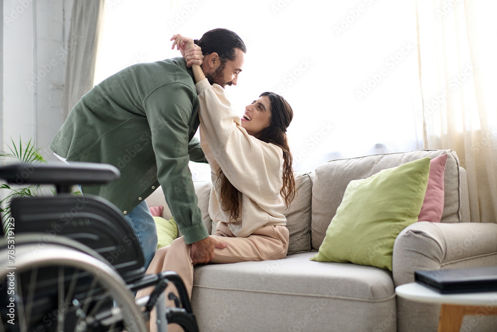 loving man in homewear helping his wife with mobility disability to get to couch from wheelchair loving man in homewear helping his wife with mobility disability to get to couch from wheelchair