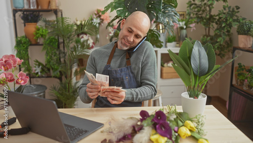 Hispanic bald man counts icelandic krona in a green plant-filled flower shop while talking on the phone. photo