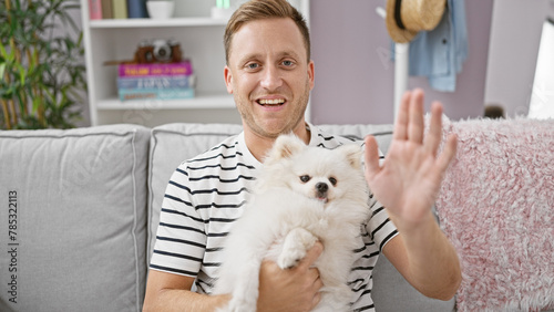 Cheerful young caucasian man happily lounging at home on his cozy sofa, confidently saying hello with a hand gesture while sitting with his joyful pet dog, both sharing a comforting smile.