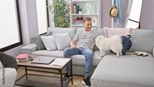 Cheerful young caucasian man confidently relaxing with his happy dog on a cozy sofa, enjoying his comfortable home interior with a joyful smile.