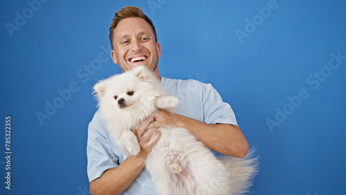 Фотография Cheerful young caucasian man laughing confidently while standing with his funny dog, radiating joy and positivity against an isolated blue wall background outdoors