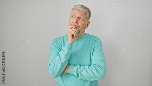 A pensive senior man with grey hair in a turquoise sweater stands against a white background, hand on chin.