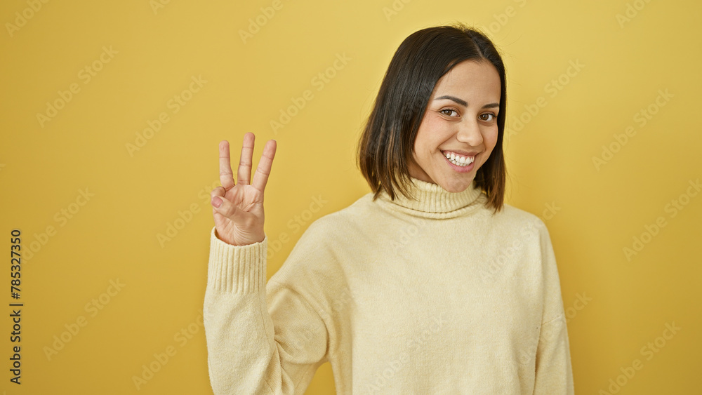 Smiling young hispanic woman gesturing number three with her fingers against a yellow isolated background. Smiling young hispanic woman gesturing number three with her fingers against a yellow isolated background.