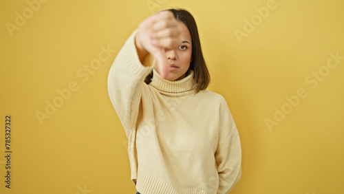 Hispanic young woman with neutral expression wearing sweater against yellow wall giving thumbs down gesture. © Krakenimages.com