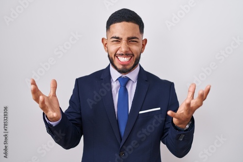 Young hispanic man wearing business suit and tie celebrating mad and crazy for success with arms raised and closed eyes screaming excited. winner concept