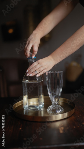 Close-up of a woman's hands unscrewing a bottle of water on a tray with an empty glass in a dark cozy restaurant setting.