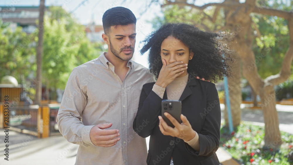 A concerned man and shocked woman reading news on a smartphone in a sunny park. A concerned man and shocked woman reading news on a smartphone in a sunny park.
