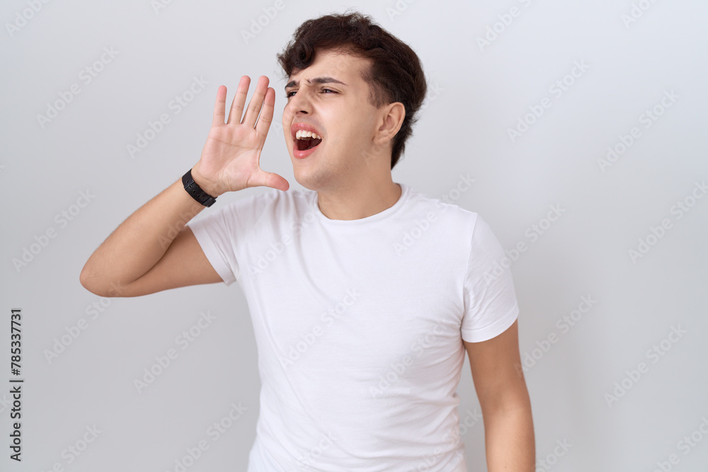 Young non binary man wearing casual white t shirt shouting and screaming loud to side with hand on mouth. communication concept. Young non binary man wearing casual white t shirt shouting and screaming loud to side with hand on mouth. communication concept.