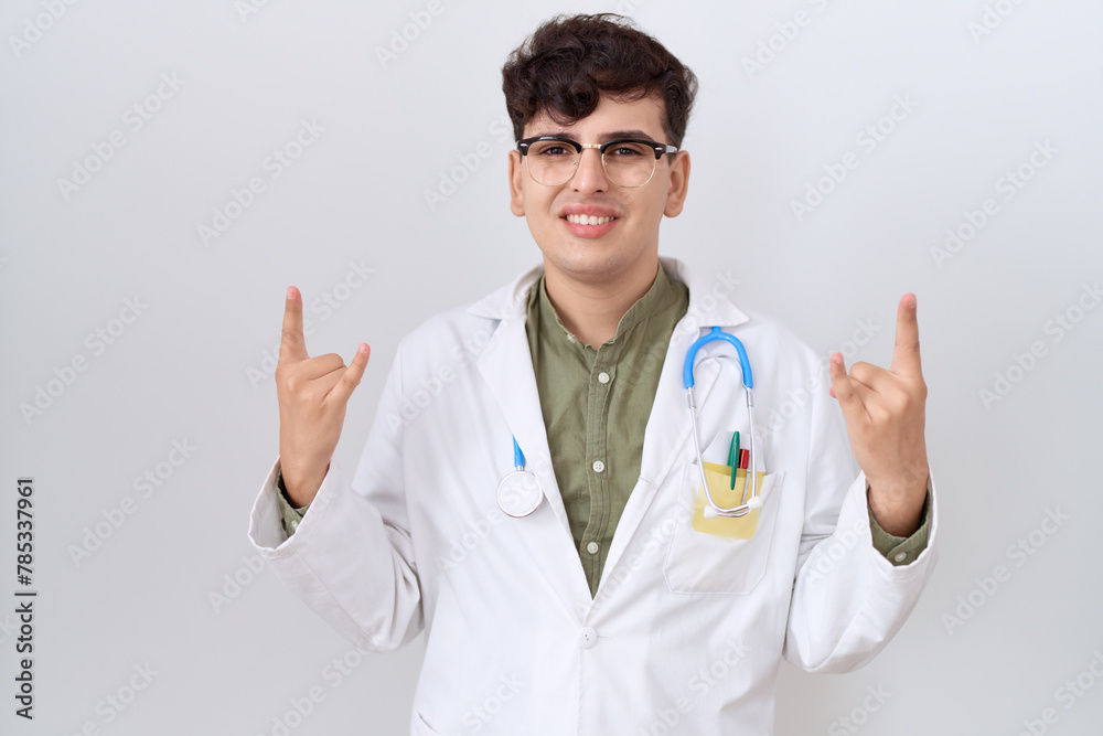Young non binary man wearing doctor uniform and stethoscope shouting with crazy expression doing rock symbol with hands up. music star. heavy concept. Young non binary man wearing doctor uniform and stethoscope shouting with crazy expression doing rock symbol with hands up. music star. heavy concept.