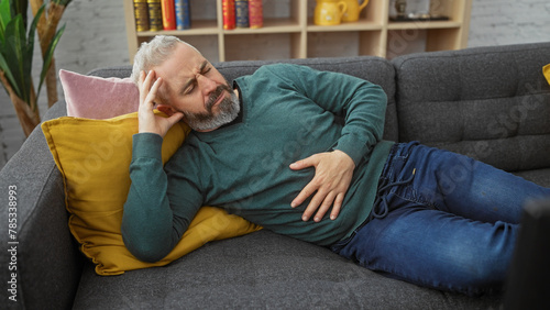 Картината върху платно A middle-aged bearded man lying on a couch at home, appearing to suffer from stomach pain