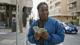 African woman in blue jacket counts money on a busy urban street, showcasing financial planning in a city setting.