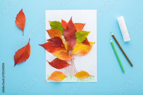 Tree shape created from colorful leaves on white paper, glue stick and pencils on light blue table background. Pastel color. Making autumn decorations. Closeup. Top view.