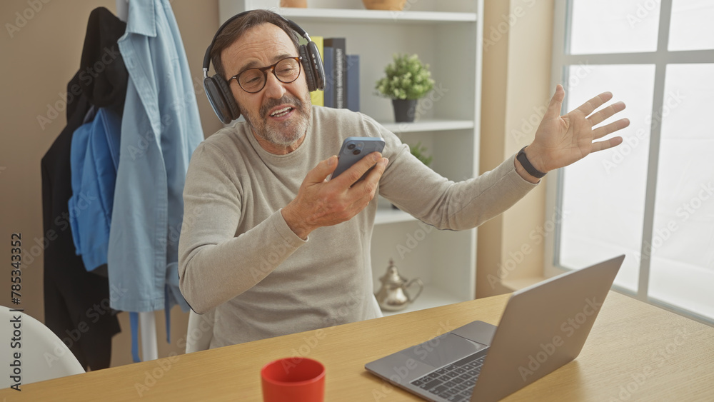 A cheerful middle-aged man with a beard video calls on a smartphone at home, with headphones and a laptop.