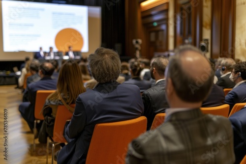 A group of business people sitting in the conference hall listening to an individual man giving a presentation on stage.