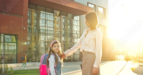 Caucasian cheerful woman holding hand of little daughter and standing near school outdoors on street. Happy girl holds mother hand in sun lights and smiling to camera. Back to school concept