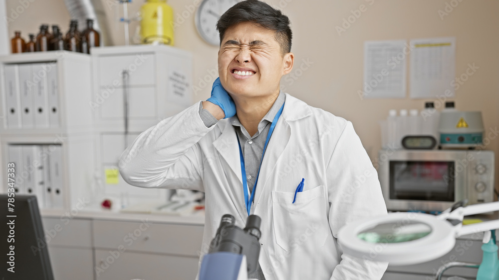 A young asian man in a lab coat grimaces, holding his neck in pain, indicating discomfort inside a modern medical laboratory.