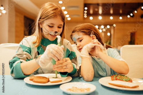Two girls with aquagrim on their faces decorate Christmas cookies with a pastry bag in a cafe or restaurant. Sisters in casual clothes have fun and paint ginger sweetness with cream. photo