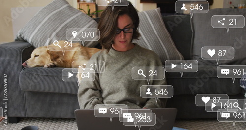 Digital icons on speech bubbles against caucasian woman using laptop while dog sleeping on couch