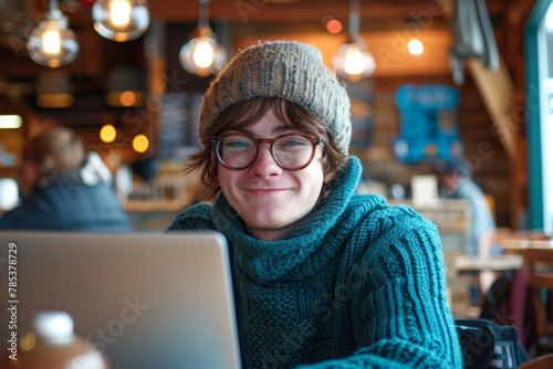 A young man wearing a blue sweater and glasses is smiling while using a laptop. He is enjoying his time at a cafe