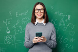 Young smiling smart teacher woman wear grey casual shirt glasses hold use mobile cell phone isolated on plain green wall chalk blackboard background studio. Education in high school college concept.
