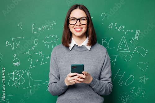 Young smiling smart teacher woman wear grey casual shirt glasses hold use mobile cell phone isolated on plain green wall chalk blackboard background studio. Education in high school college concept. © ViDi Studio