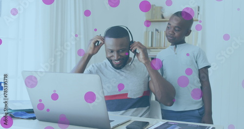 Image of pink spots floating over african american father and son using phone headset and laptop