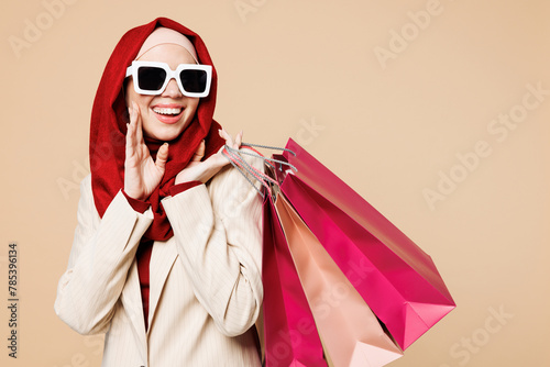 Young fun Arabian Asian Muslim woman wear red abaya hijab suit clothes shopping hold package bags look aside isolated on plain beige background. Black Friday sale buy day, UAE Islam religious concept.