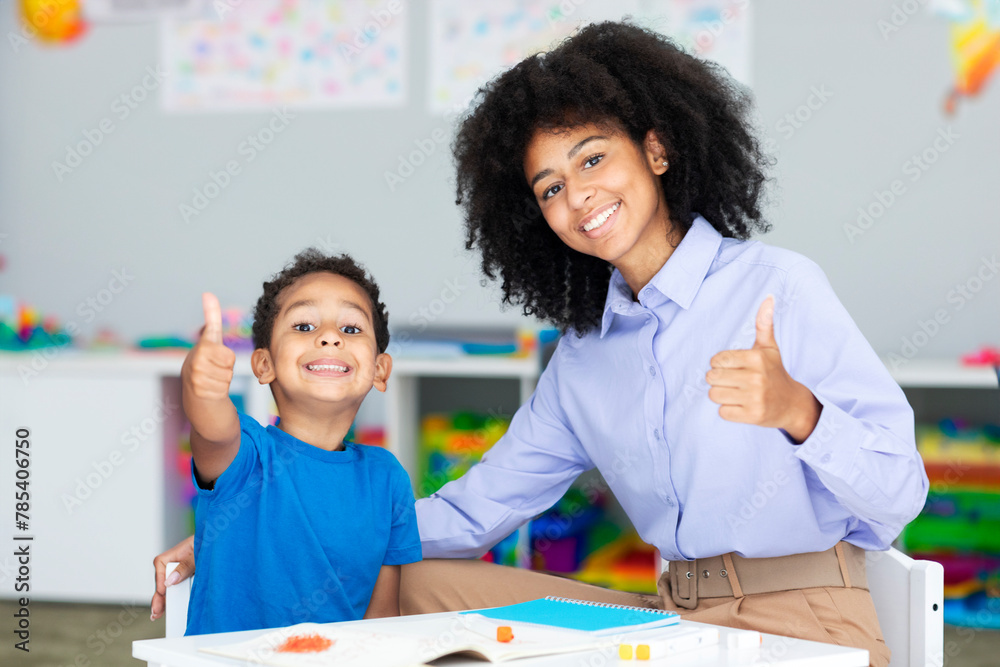 Tutoring and mentorship concept. Black female teacher and happy schoolboy gesturing thumbs up and smiling sitting at desk in class Tutoring and mentorship concept. Black female teacher and happy schoolboy gesturing thumbs up and smiling sitting at desk in class