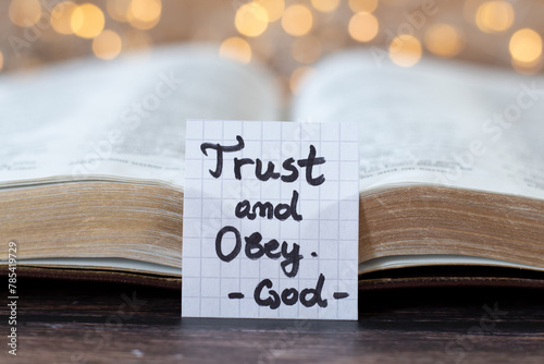 Trust and obey, God, handwritten quote with open holy bible and bokeh light background. Close-up. Faith and belief in Jesus Christ, Christian obedience, biblical concept.