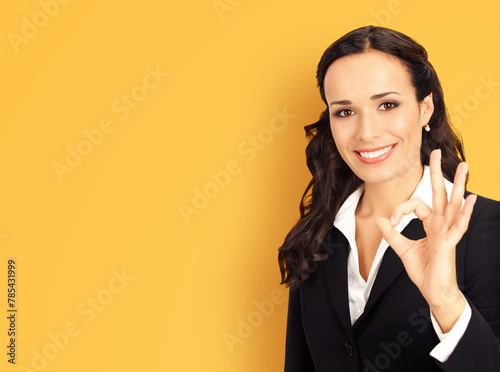 Happy smiling brunette businesswoman business woman wear black confident suit, show okay hand sign gesture, isolated against yellow wall background. Blank empty free ad text area.