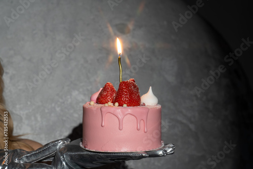 girl hand holds glamorous pink strawberry birthday cake specially for woman with burning candle on it. Make a wish, blow a candle. Starlight effect on glowing flame, blazing star. Celebration day