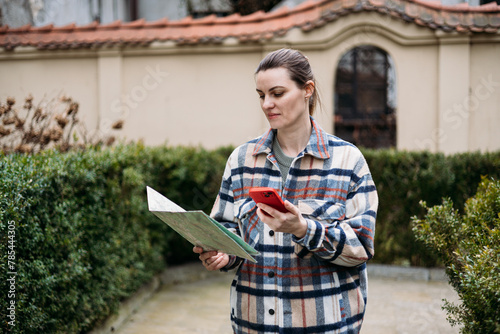 Attractive young female tourist is exploring city. Happy woman holding a paper map and phone on city street. Traveling Europe in autumn, urban background