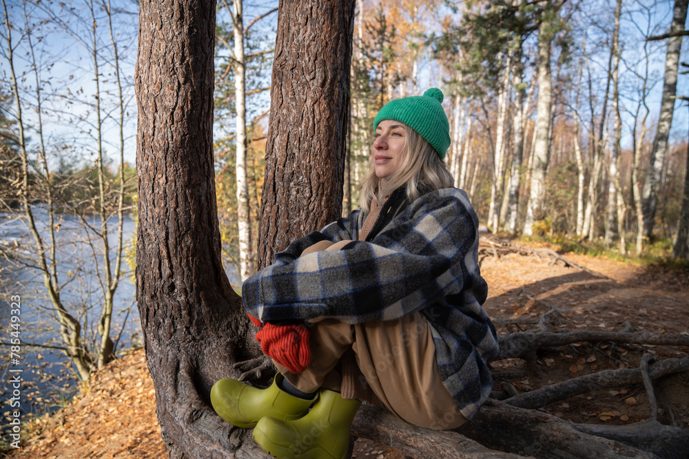 Middle aged woman contemplating enjoying nature in park sitting on tree roots. Relaxed female in warm clothes resting in autumn forest on lake shore. Slow life, retreat concept Middle aged woman contemplating enjoying nature in park sitting on tree roots. Relaxed female in warm clothes resting in autumn forest on lake shore. Slow life, retreat concept