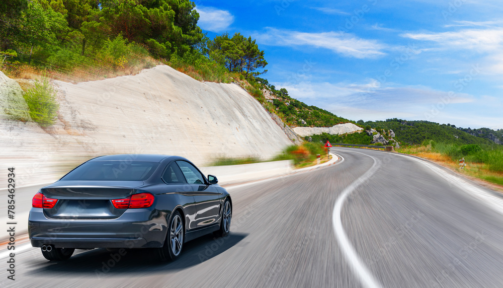 A white car drives along the highway against the backdrop of rocky mountains on a sunny day.