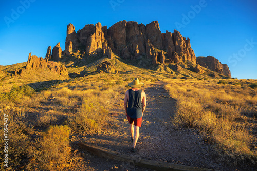 Hiker in a straw hat walking towards rock formations named Superstition Mountains in Lost Dutchman State Park, Arizona.