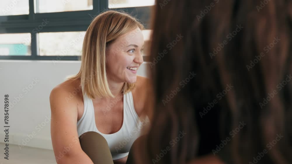 A smiling adult woman in sportswear sits and talks to fellow participants before starting a stretching class in a studio setting.