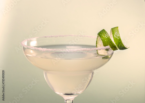 Close up view of classic margarita cocktail garnished with salt rim and lime spiral zest on subtle pastel green color background