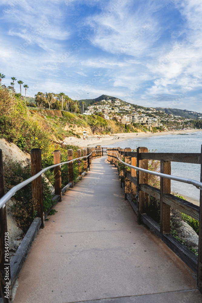 A wooden boardwalk leads to the beach. The boardwalk is lined with trees and has a railing. The beach is a beautiful, serene place with the ocean in the background