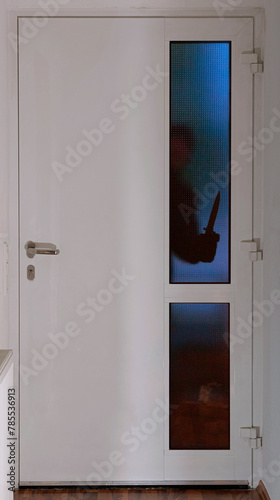 Ein Einbrecher steht in der Dunkelheit mit einem Messer vor einer Eingangstüre und schaut durch die Glasscheibe in die Wohnung oder Haus rein

