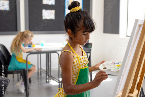 In school, during art class, a young biracial girl with dark hair is holding a paint palette, painti
