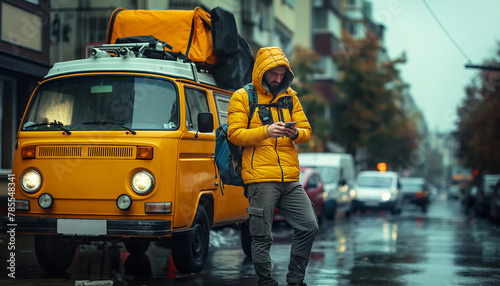 Man in yellow jacket stands next yellow camper van on rainy urban street, checking his phone. Image depicts traveler navigating through city in inclement weather. Tarvel in campers, navigation concept photo