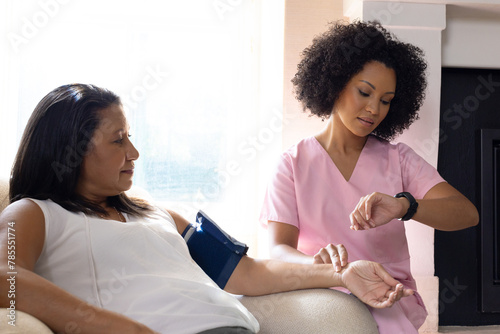Mature biracial woman checking blood pressure at home, young nurse assisting